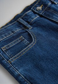 Közelkép kék farmernadrág elülső zsebéből, fém gombbal, övbújtatóval és narancssárga varrási részletekkel.