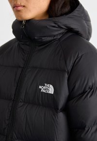 Crna izolirana puffer jakna s kapuljačom, koja ima puni patentni zatvarač i vidljiv logo na prsima. Glatka tekstura i kvadratni dizajn.