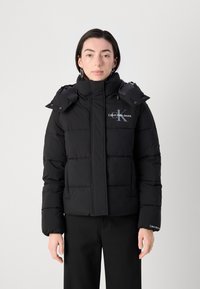 Zwarte pufferjack met een hoge kraag en afneembare capuchon, voorzien van gestikte delen en een "Calvin Klein Jeans" logo op de borst.