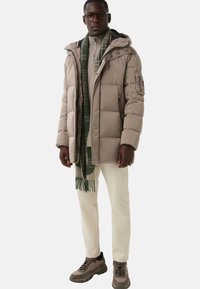 Puffer jacket in light beige, featuring a hood and zip pockets. Paired with a green plaid scarf, cream pants, and gray-brown hiking shoes.