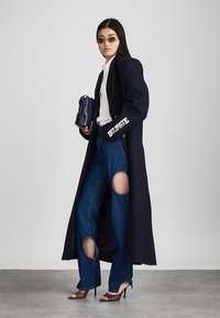 Μακρύ ναυρίνιο παλτό με υπερμεγέθη εφαρμογή, λεπτομέρεια λευκού λογότυπου στα μανίκια, λευκό πουκάμισο, σκισμένο τζιν με μεγάλες ανοιχτές περιοχές, καφέ ψηλοτάκουνες μύτες, γυαλιά ηλίου.