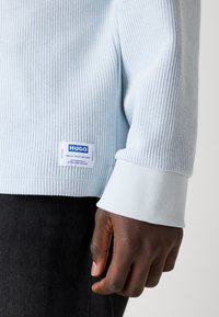 Hanorac ribbed de culoare albastru deschis, cu manșete albe; are un etichetă cu "HUGO" în culoare albastră. Materialul pare moale și texturat.