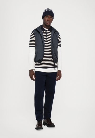 Mann steht da, trägt ein gestreiftes langärmliges Hemd, eine schwarze Steppweste, eine marineblaue Hose, dunkelbraune Schuhe und einen dunklen Strickhut vor einem schlichten Hintergrund.