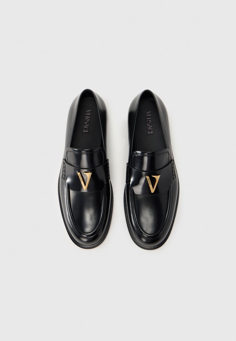 Mocassins en cuir noir à finition brillante, avec un ornement en V doré proéminent et un design d'orteil lisse et arrondi.