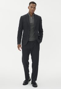 Antracytowy garnitur w kratę z dopasowaną marynarką i plisowanymi spodniami, połączony z szarym topem z zamkiem na kołnierzu oraz czarnymi butami na sznurówki.