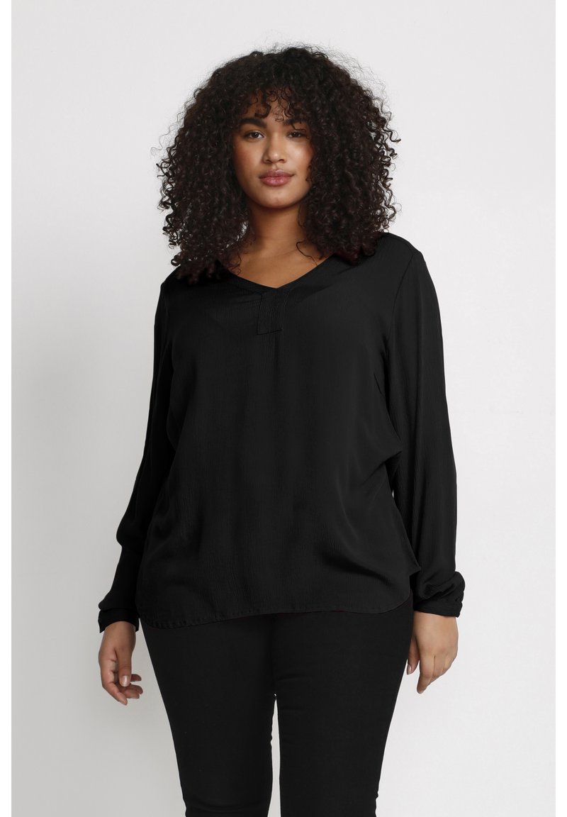 Blouse noire à manches longues avec un décolleté en V et une coupe décontractée, présentant un tissu texturé et des poignets de manches froncés. Portée avec un pantalon noir.