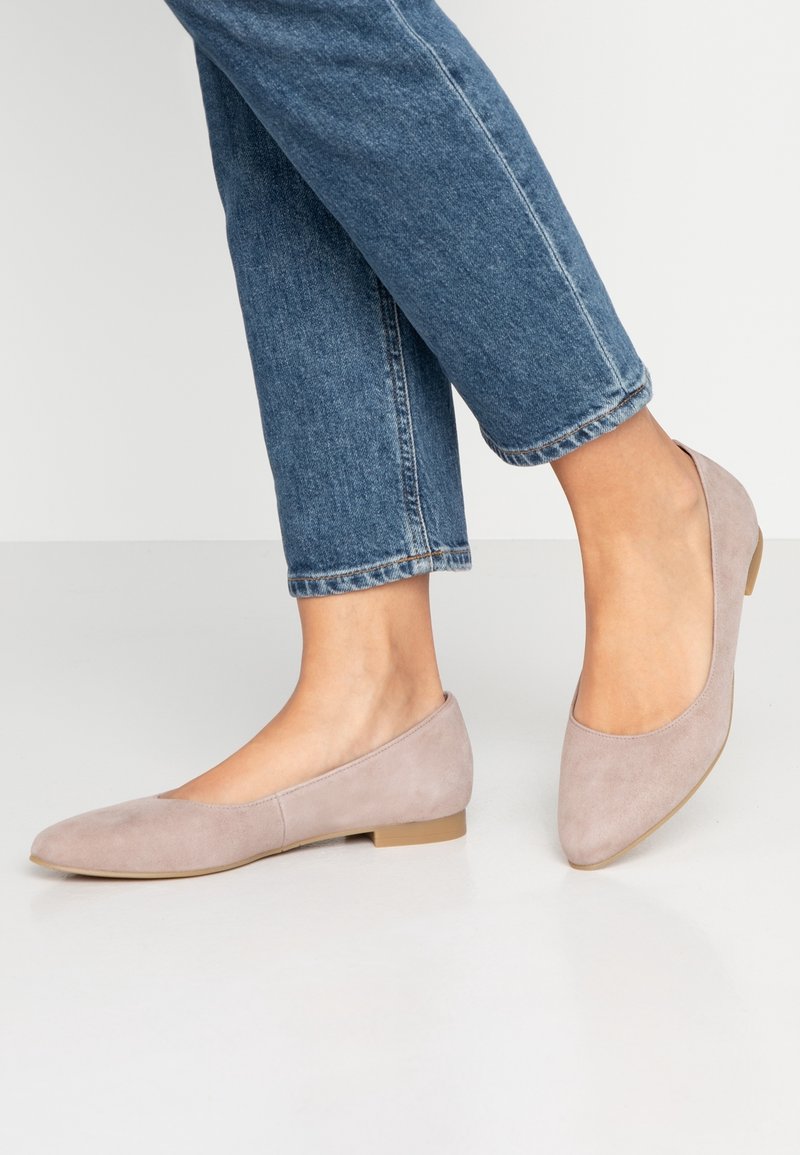 Pieds portant des ballerines en daim rose clair associées à un jean bleu en denim sur un fond clair uni.