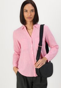 Light pink button-down shirt made of textured fabric, featuring a chest pocket and long sleeves, paired with a black crossbody bag.