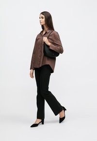 Femme marchant, portant une chemise marron à boutons, un pantalon noir, des talons noirs, et portant un petit sac à main noir sur fond blanc.