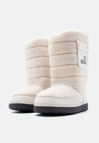 Χειμερινές μπότες από απαλό, μπεζ φλις με μαύρες σόλες, στρογγυλεμένες μύτες και ένα λευκό έμβλημα με την επιγραφή "LOVE MOSCHINO" στο πλάι.