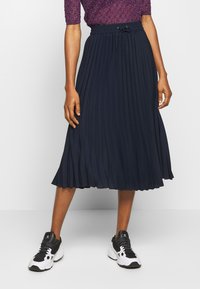 Gonna plissettata navy con vita elastica e cordino. Indossata con sneakers da ginnastica nere e bianche. Il tessuto testurizzato aggiunge interesse visivo.