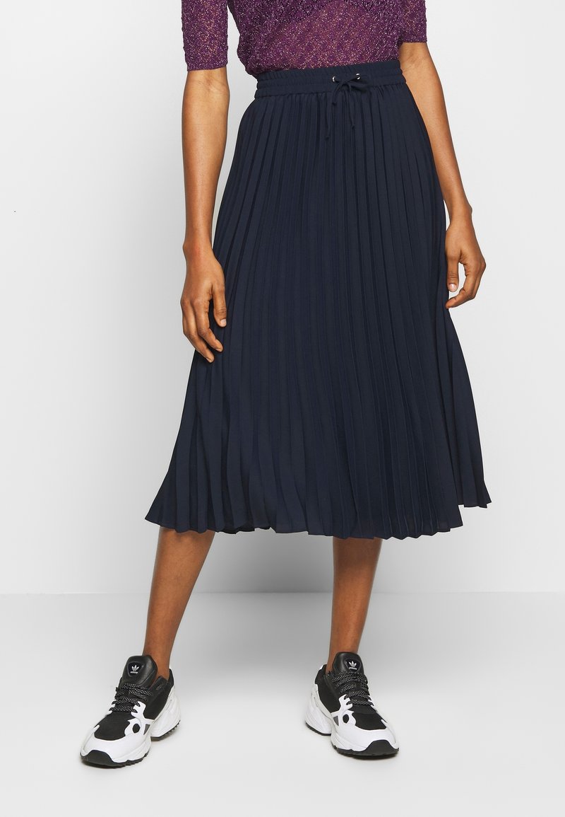 Gonna plissettata navy con vita elastica e cordino. Indossata con sneakers da ginnastica nere e bianche. Il tessuto testurizzato aggiunge interesse visivo.