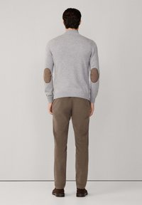Hombre de pie de espaldas, vistiendo un suéter gris claro con parches marrones en los codos, pantalones marrones y zapatos marrones oscuros sobre un fondo liso.