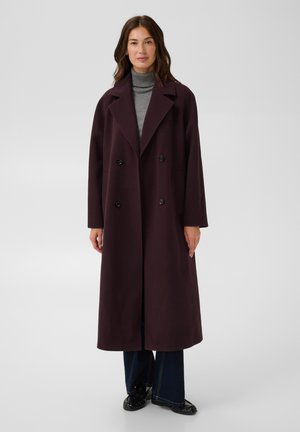 Femme debout portant un long manteau violet foncé à double boutonnage, un col roulé gris, un pantalon large foncé et des chaussures noires sur un fond uni.