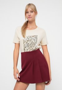 Beige t-shirt met een textuur van een luipaardprint, gecombineerd met een bordeauxrode geplooide rok. Soepele stof met een aangesloten taille.