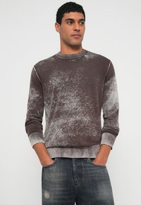 Mann mit kurzen lockigen Haaren, der einen braun-grau verwaschenen Langarmpullover und zerrissene blaue Jeans trägt, die Hände in den Taschen, schaut zur Seite.