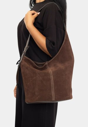 Mujer con un atuendo negro que lleva un gran bolso de hombro de ante marrón con costuras visibles en el hombro izquierdo.