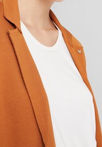Blazer marrone con texture e colletto, indossato sopra una maglietta bianca a collo tondo, con un piccolo accento metallico sul blazer.