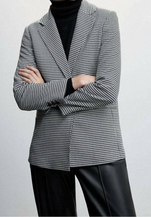 Personne croisant les bras portant un blazer pied-de-poule noir et blanc, un col roulé noir et un pantalon en cuir noir devant un fond uni.