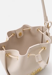 Beige faux läder bucket bag med gyllene detaljer, snörstängning och mjukt inre, med gyllene accenter på utsidan.