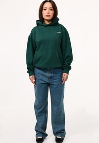 Grüner Baumwoll-Hoodie mit Kängurutasche und Kapuze mit Kordelzug, kombiniert mit hellblauen geraden Jeans und schwarzen Schuhen.