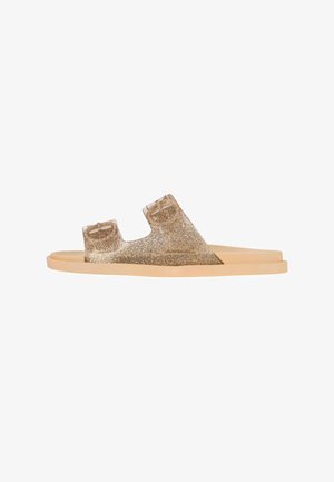 Infradito glitter dorate con due cinture larghe, suola piatta color tan e materiale superiore trasparente che mette in mostra dettagli scintillanti.
