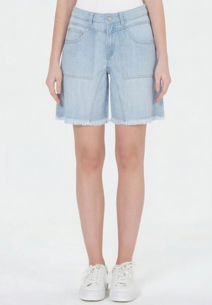 Hellblaue Denim-Shorts mit aufgesetzten Fronttaschen, ausgefranstem Saum, getragen mit weißen Sneakers und weißem Oberteil, stehend vor einem schlichten Hintergrund.