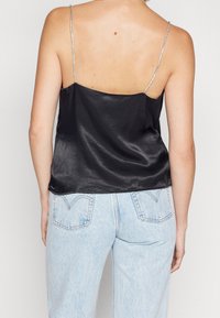 Camisole en satin noir avec fines bretelles spaghetti argentées et texture lisse, portée avec un jean en denim bleu clair doté de poches arrière.