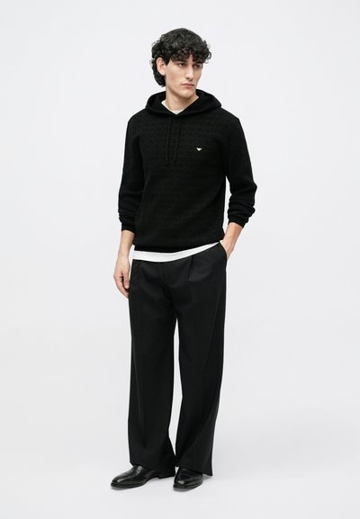 Camisola preta texturizada com capuz e logótipo dourado, sobreposta a uma camisa branca, combinada com calças pretas de perna larga e sapatos pretos.