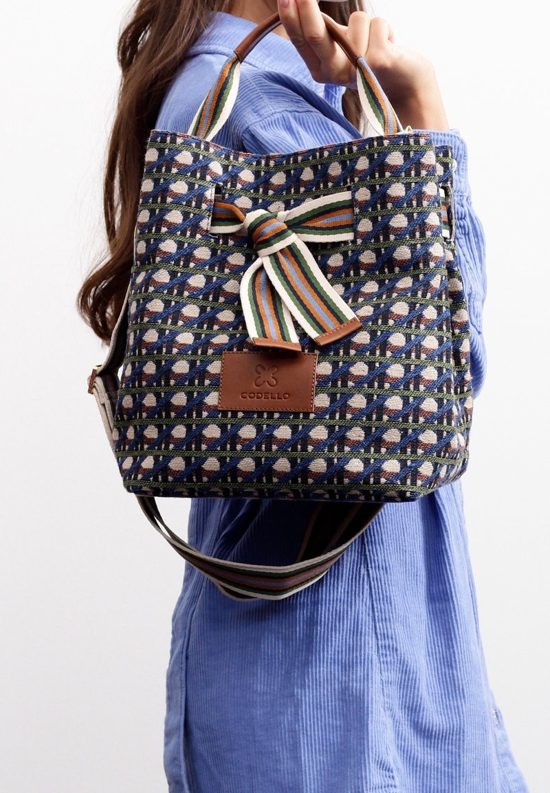 Niebieska torba z wzorzystej tkaniny z brązową skórzaną metką. Posiada paskowany uchwyt z tkaniny i ozdobę w formie kokardki. Prostokątny kształt i średni rozmiar.