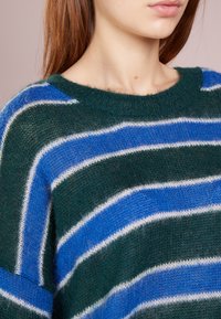 Groen gebreide trui met blauw-witte horizontale strepen, voorzien van een ronde hals en een zachte textuur. Close-up van het stofpatroon.