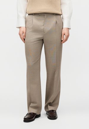 Bézs, szabott nadrág, fehér inggel és bézs kötött mellénnyel viselve, sötétbarna bőr mokaszinokkal, fehér háttér előtt.