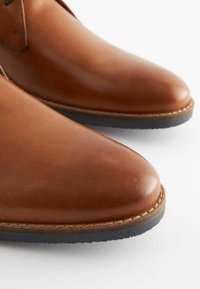 Zapatos de cuero marrón con una textura suave, puntera afilada y suela cosida en contraste. Presentan un diseño minimalista con un brillo sutil.