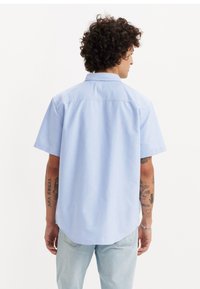  kortærmet lyseblå skjorte med buet kant, knapkrave og glat stof; båret med slidte jeans og synlige tatoveringer på armene.