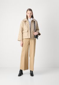 Beige quilted jacket with a collared neckline, front pocket, paired with wide-legged tan trousers and black heeled ankle boots.