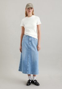 Weißes, eng anliegendes T-Shirt, hellblaue Jeans-Maxiröhre, schwarze Loafer mit goldenen Akzenten und hellfarbige Socken. Olivgrüne Mütze mit Logo.