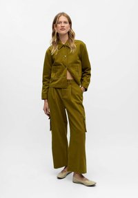 Veste courte de couleur olive avec deux poches avant, associée à un pantalon large avec des poches cargo sur les côtés. Le modèle porte des chaussures plates beiges.