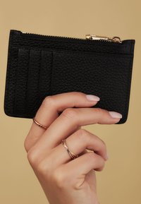 Black textured cardholder with multiple slots and a zip closure, featuring gold hardware accents. Held between fingers.