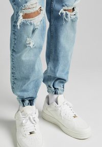 Ljust blå slitna denimjoggare i kombination med vita sneakers och vita strumpor på en neutral bakgrund.