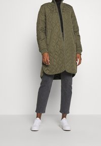 Chaqueta larga acolchada de color verde oliva con cremallera frontal, puños y dobladillo acanalados, llevada sobre una camiseta negra y jeans grises, combinada con zapatillas blancas.