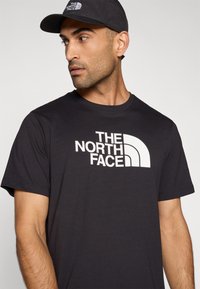 Μαύρο βαμβακερό T-shirt με το λογότυπο "THE NORTH FACE" σε λευκό, στρογγυλή λαιμόκοψη και κοντά μανίκια. Απλός σχεδιασμός με ελάχιστες λεπτομέρειες.