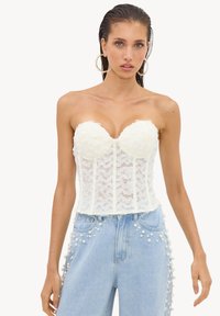 Femme portant un corset bustier en dentelle blanche et un jean bleu clair avec des ornements perle le long des poches et des côtés.