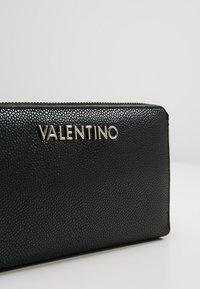 Portefeuille en cuir noir texturé avec un logo "VALENTINO" en métal argenté, doté d'une fermeture éclair et d'un design rectangulaire.