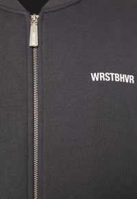 Schwarze Zip-Hoodie aus geripptem Stoff, mit einem silbernen Reißverschluss und weißem "WRSTBHV"-Logo auf der linken Brust. Glatte Textur.
