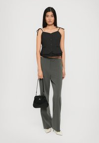 Woman wearing black buttoned sleeveless top, gray pinstripe wide-leg pants, white heels, holding black handbag, standing against white background.