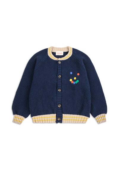 Mornarska pletena jakna ima rebrasti žuti i bež ovratnik i manžete, pet dugmadi i šarena točkasta uzorka na prsima.