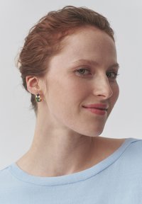 Pendiente de aro dorado y colorido llevado por una persona con cabello rojo, con superficies lisas y un diseño moderno y elegante. Parte superior azul clara visible.