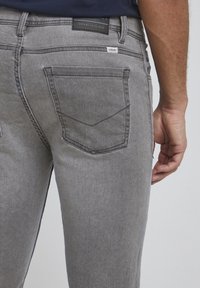Szare dżinsy denim o wąskim kroju, z tylnymi kieszeniami z przeszyciami oraz metką na pasku. Gładka faktura, casualowy design.