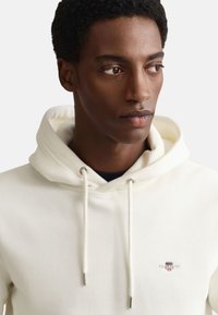 White hooded sweatshirt with drawstrings, silver hardware, and a small logo patch on the chest. Smooth fabric, casual design.