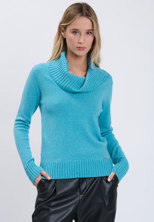 Junge Frau in hellblauem Rollkragenpullover und schwarzen Lederhosen steht mit den Händen in den Taschen vor einem schlichten Hintergrund.